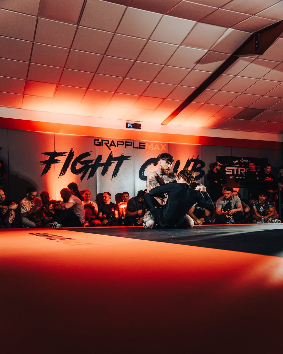 Two competitors grappling on the mats at GrappleMax Fight Club under red lighting